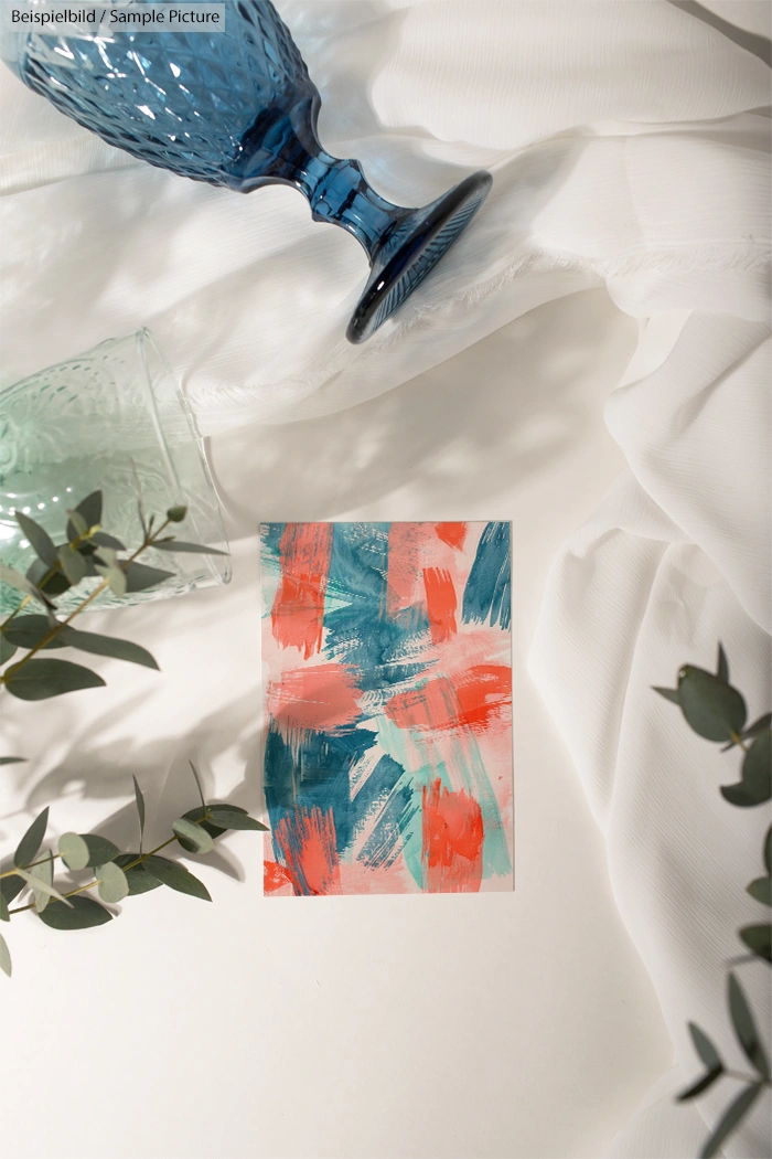 Colorful abstract painting beside glass vases and greenery on a white cloth surface.