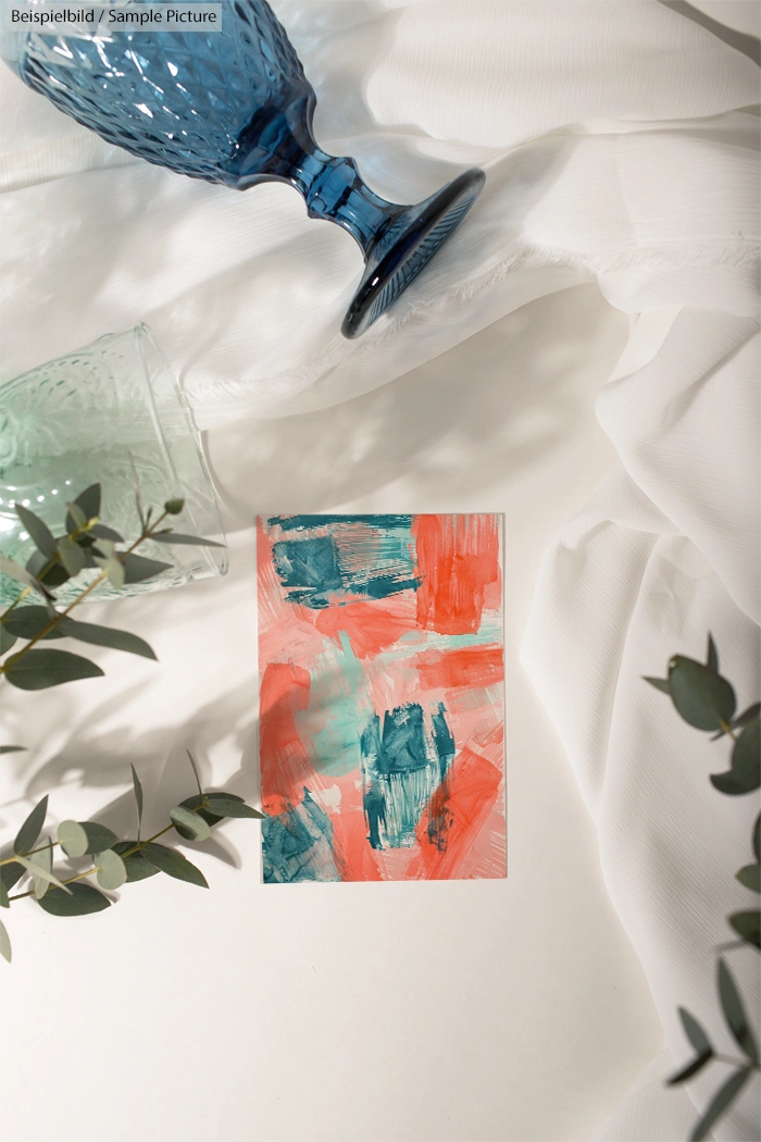 Abstract painting with blue and coral strokes on a white surface, next to blue glassware and greenery.