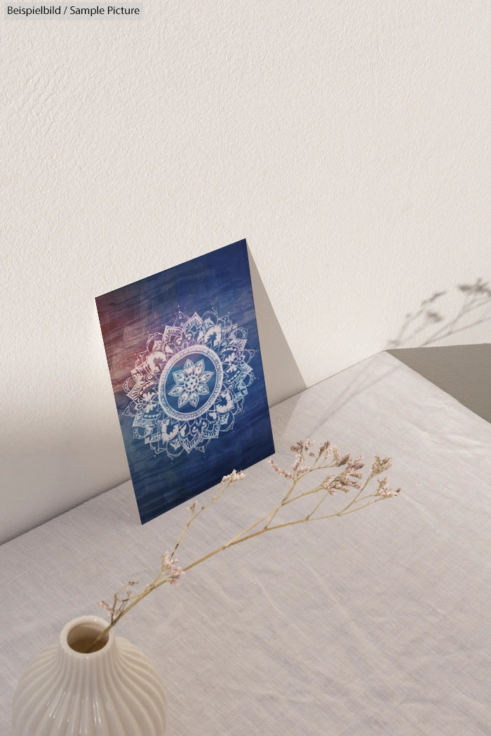 Postcard with white floral mandala on blue background, standing on a white textured surface with a small vase in the foreground.