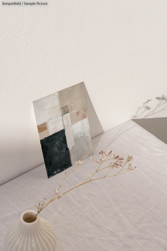 Abstract painting with neutral tones on a white table, next to a vase with delicate dried flowers.