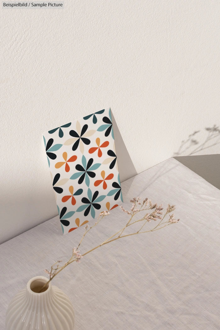 Greeting card with colorful floral pattern on a white table next to a beige vase with dried flowers.