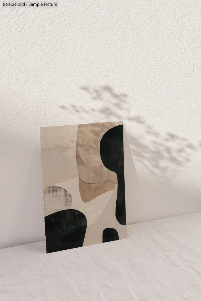 Abstract artwork on canvas with beige, black, and gray shapes, casting a shadow on a textured white wall.