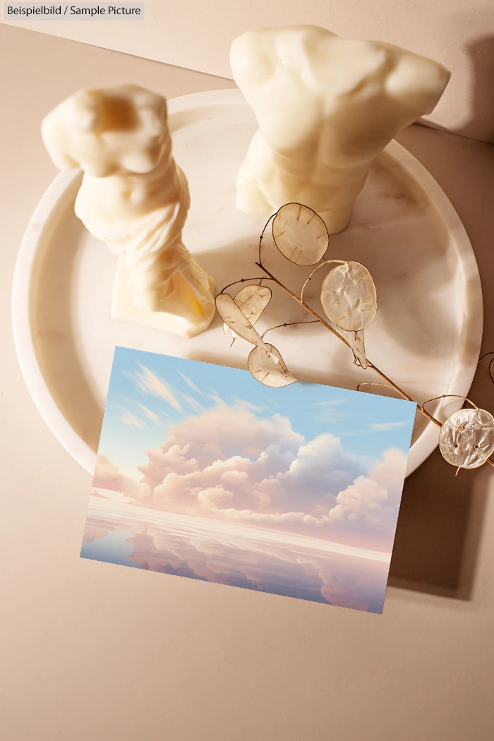 Decorative marble statues and delicate plant next to serene sky painting on a light-colored surface.