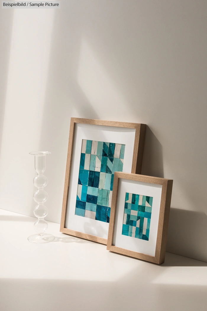 Two framed geometric artworks with blue and turquoise patterns on a light surface next to a glass candle holder.