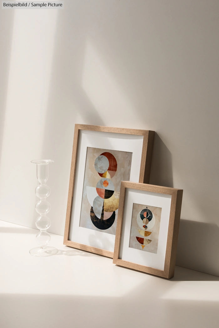 Two modern abstract art prints in wooden frames with a clear glass vase on a sunlit white surface.