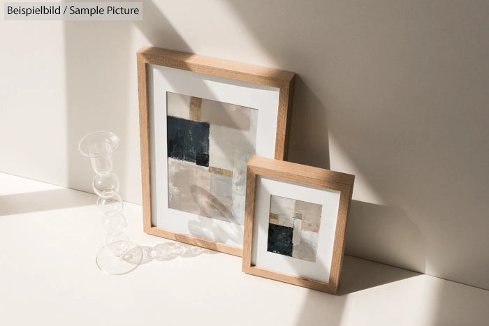 Framed abstract paintings with beige and black tones, placed on a sunlit surface next to a decorative glass object.