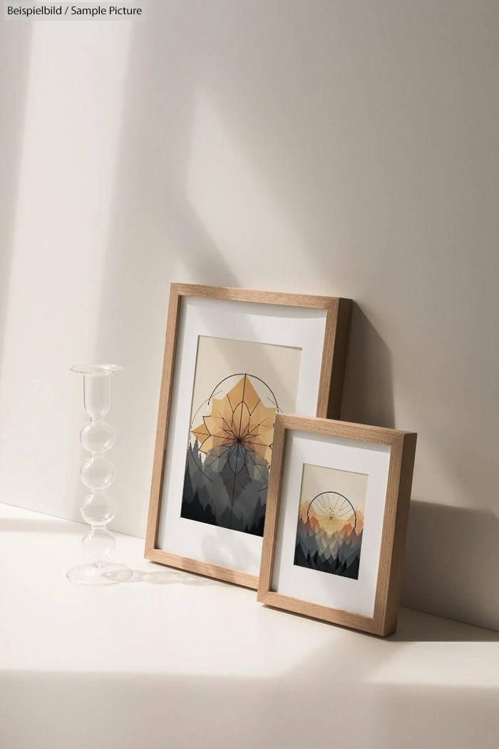 Two framed landscape prints with geometric designs, next to a clear glass candle holder on a sunlit surface.