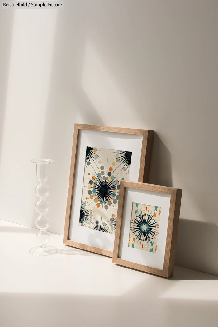 Two framed abstract paintings with geometric designs beside a clear glass vase on a sunlit surface.