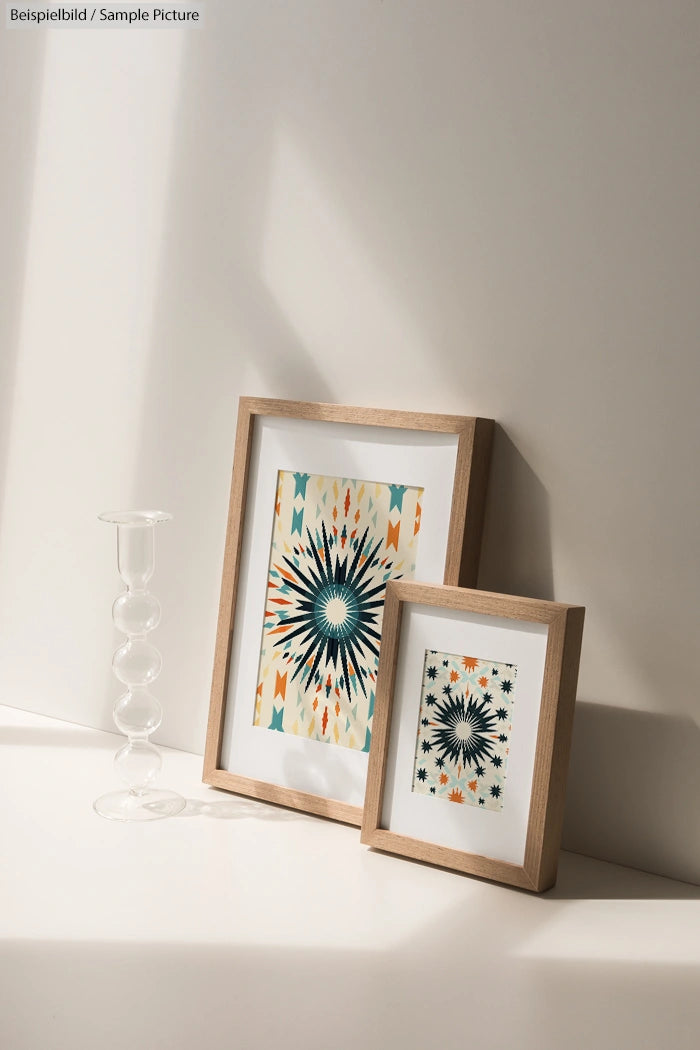 Two framed geometric prints with star patterns on a sunlit surface, beside a clear glass candle holder.