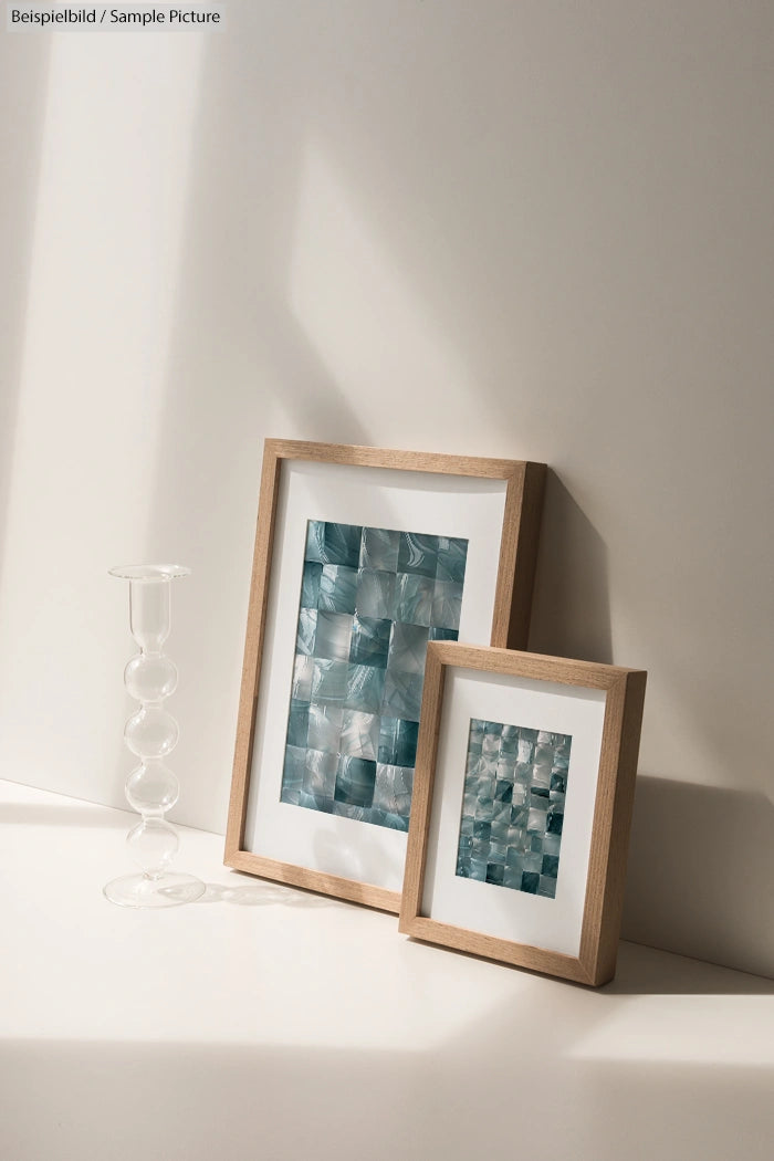 Two wooden picture frames with abstract blue mosaic patterns, placed on a white surface next to a clear glass candle holder.