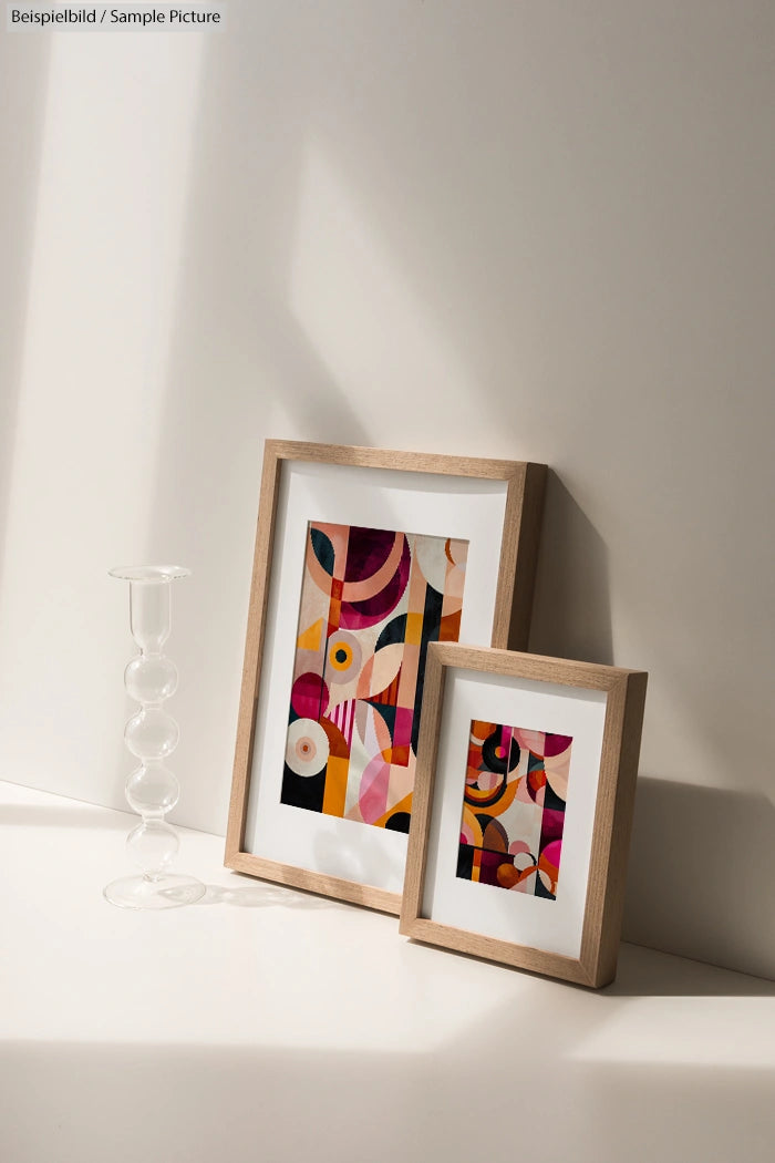 Two framed abstract artworks with geometric shapes in warm colors, placed on a sunlit surface next to a clear glass vase.