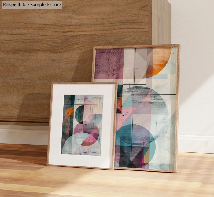 Two framed abstract artworks with geometric patterns and colorful shapes, leaning against a wooden surface.