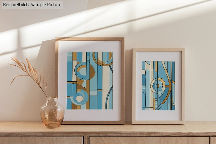 Two framed abstract artworks with blue, beige, and brown geometric patterns on a wood surface under soft lighting.