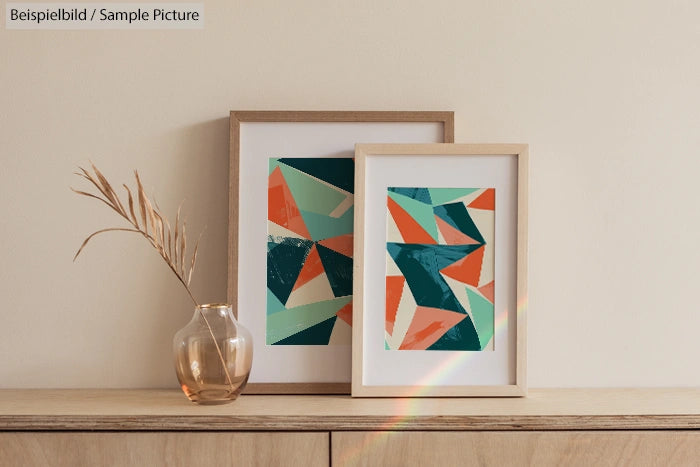 Two framed abstract paintings with geometric patterns displayed on a wooden table beside a glass vase with dried plant.