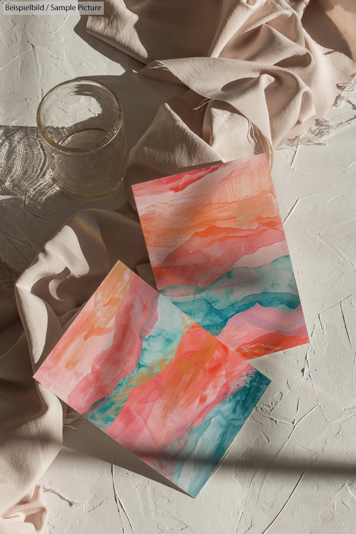 Two watercolor paintings with pink, blue, and orange tones on textured surface, partially covered by a beige cloth.