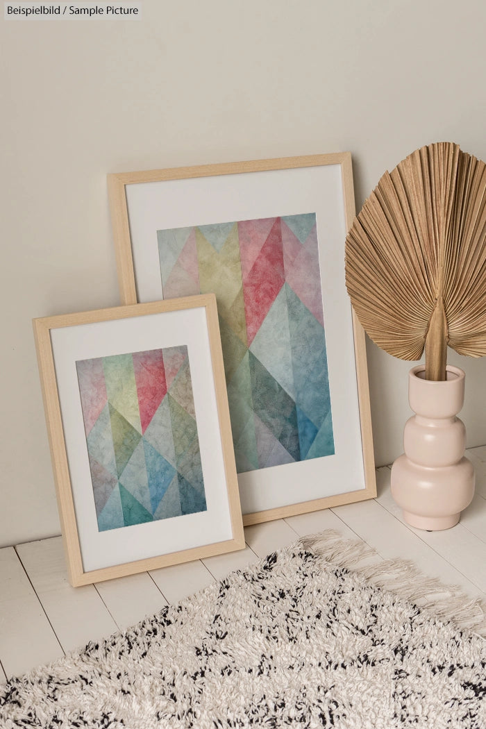 Two framed geometric artworks with pastel colors leaning against a wall, next to a large dried leaf in a pink vase.