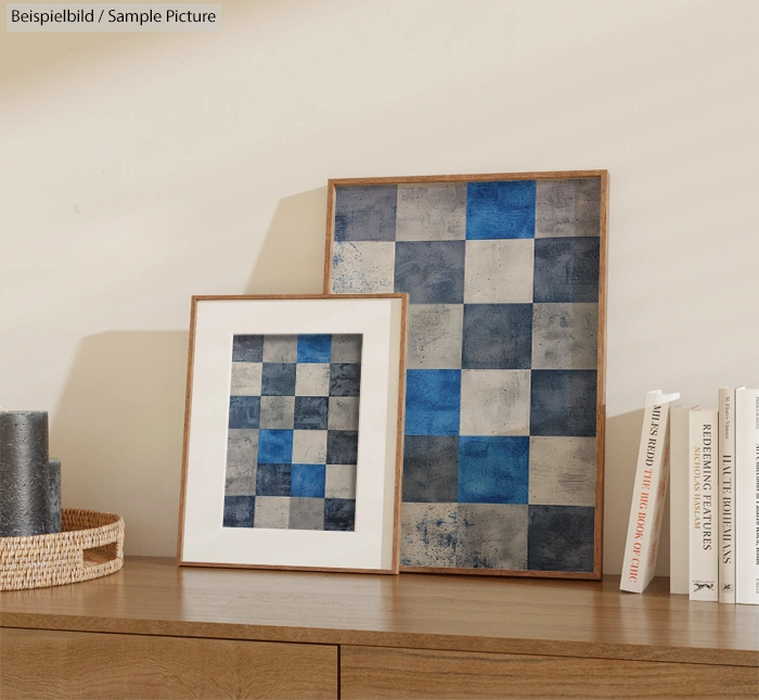 Two framed abstract paintings with blue and gray checkered patterns on a wooden shelf beside books and a basket.