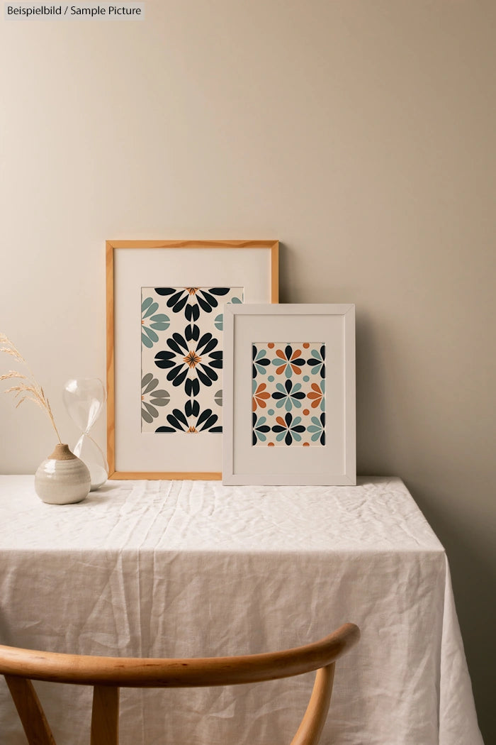 Two framed floral pattern prints on a cloth-covered table with a wooden chair, vase, and hourglass.