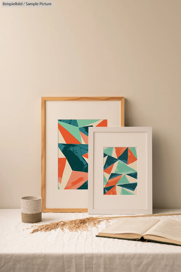 Two abstract geometric paintings in wooden frames on a table with an open book and a small plant.