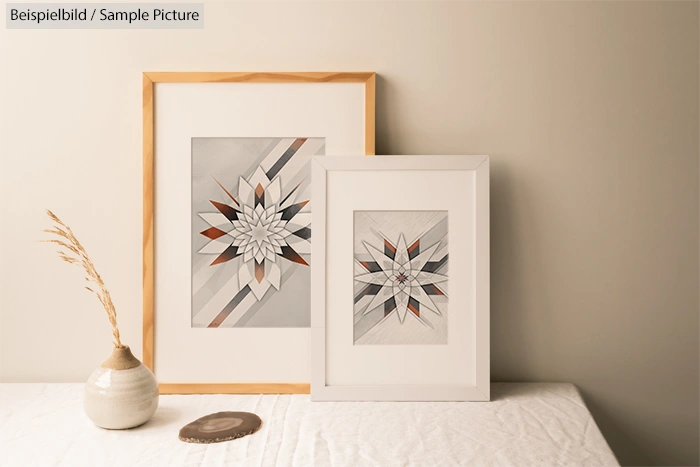 Two framed geometric art prints on a table next to a vase with dried grass against a beige wall.