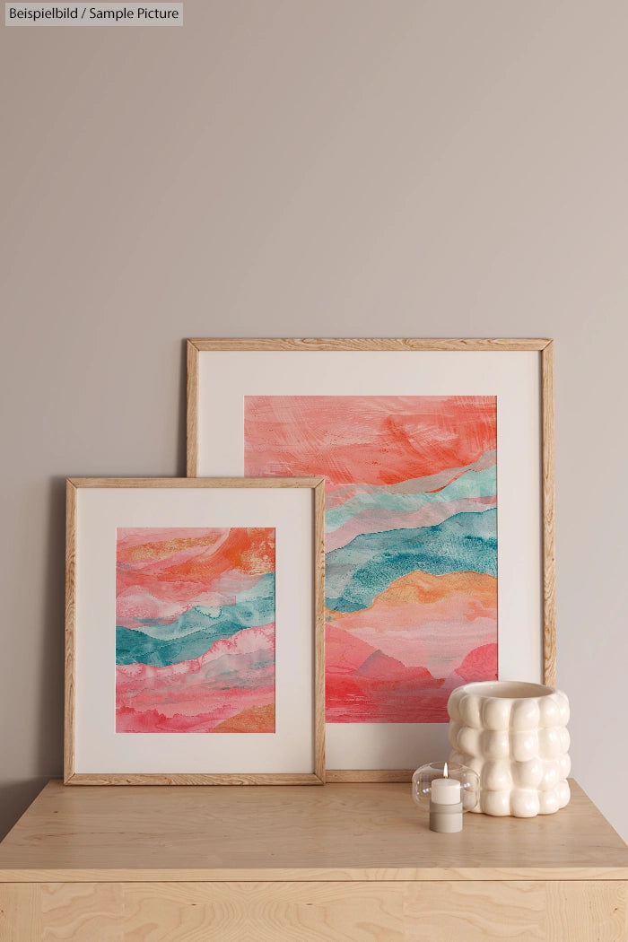 Two framed abstract paintings with coral, blue, and pink swirls on a wooden surface beside a textured white vase.