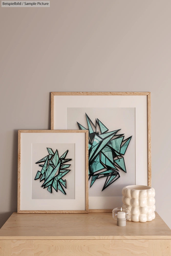 Two framed geometric artworks on a wooden surface beside a ceramic vase with a small candle.