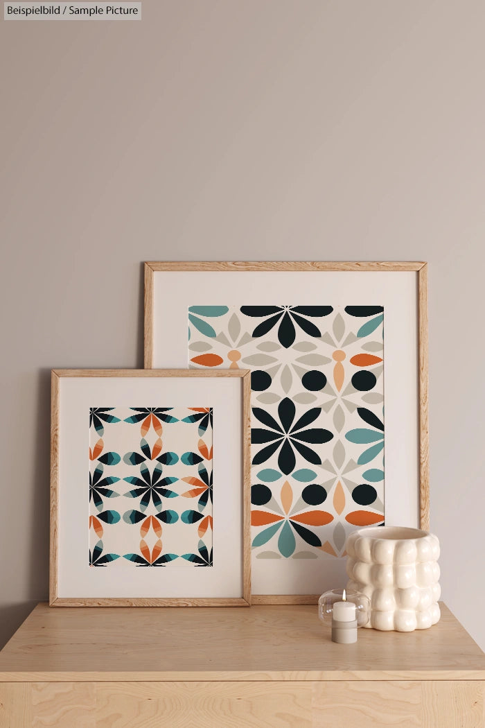 Two framed abstract prints with geometric patterns in orange, teal, and black on a wooden table with ceramic vases.