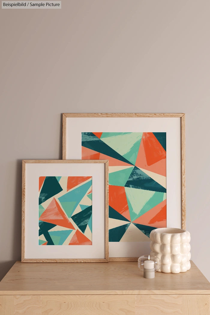 Two framed geometric abstract artworks with colorful triangles, displayed on a wooden shelf next to a textured white vase.