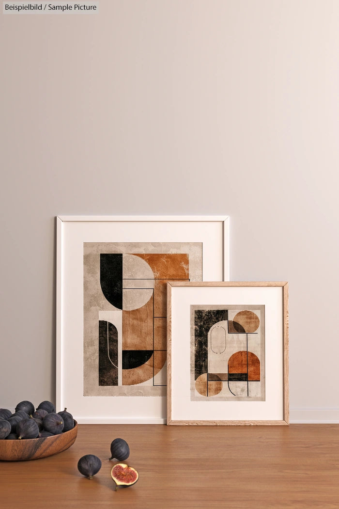 Two framed abstract art pieces on a wooden surface with a bowl of figs nearby.