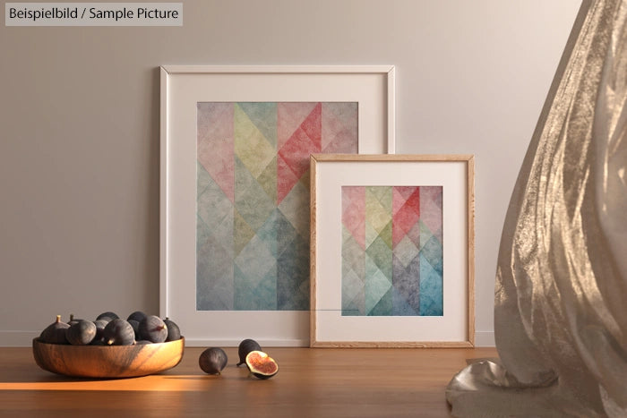 Geometric multicolored artwork in frames beside a bowl of figs, with sunlight casting shadows on a wooden surface.