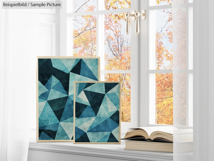 Two abstract geometric paintings in blue on a windowsill, with autumn trees visible outside the window.