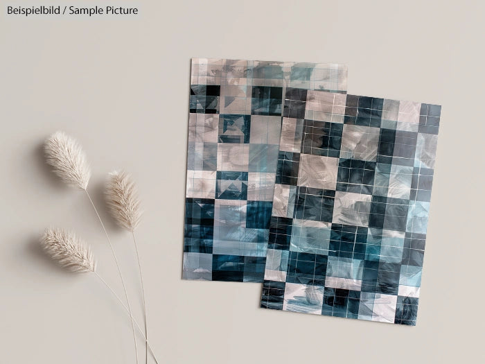 Two abstract art pieces with blue and grey checkered patterns on a light background next to dried flowers.