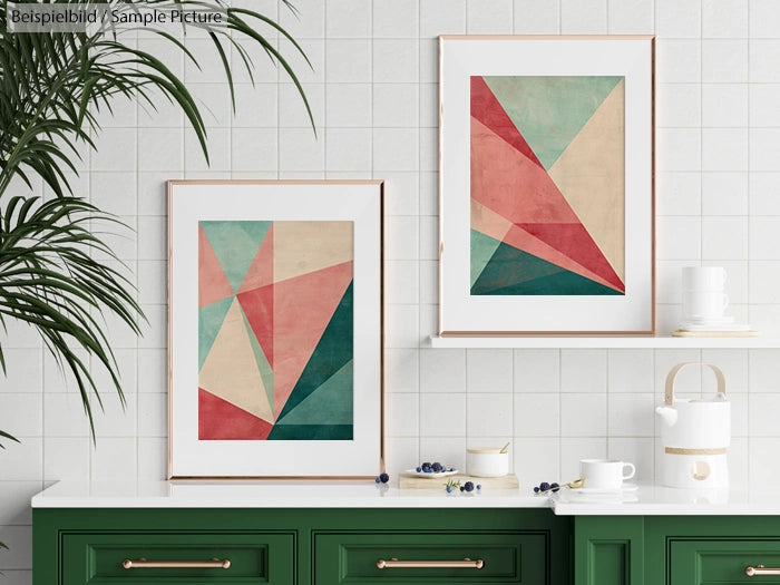 Two geometric abstract artworks displayed above a green cabinet in a minimalist white-tiled kitchen.