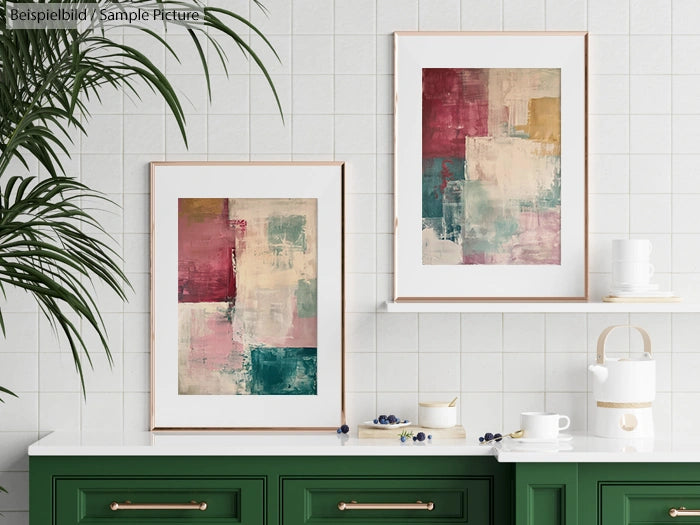 Two framed abstract paintings on white tiled wall above a green cabinet with plants and white decor.