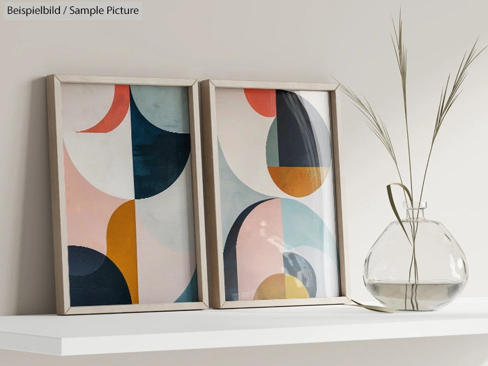 Two framed abstract art pieces on a shelf with a glass vase and grass stems.