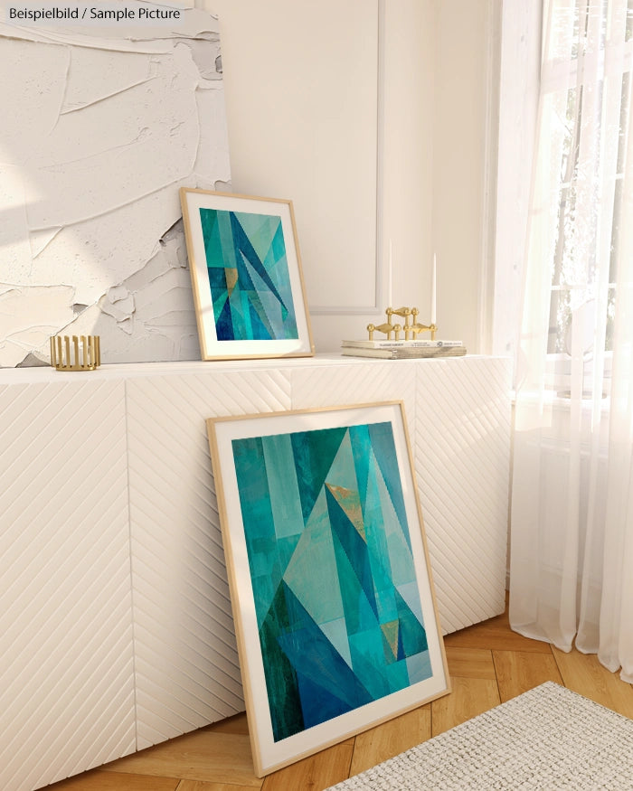 Frames with abstract blue and teal art leaning against a white cabinet in a sunlit room.