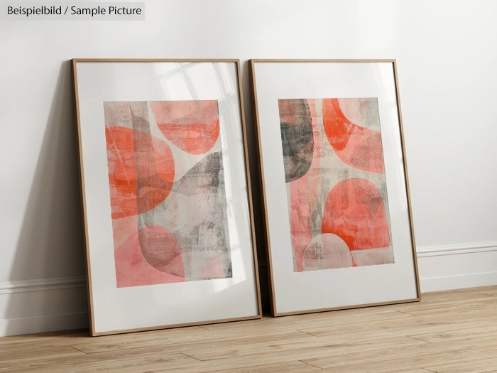 Two abstract prints with pink and gray organic shapes in wooden frames leaned against a white wall on a wooden floor.