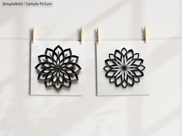 Two geometric floral designs on paper, clipped on a string, casting shadows on a plain background.