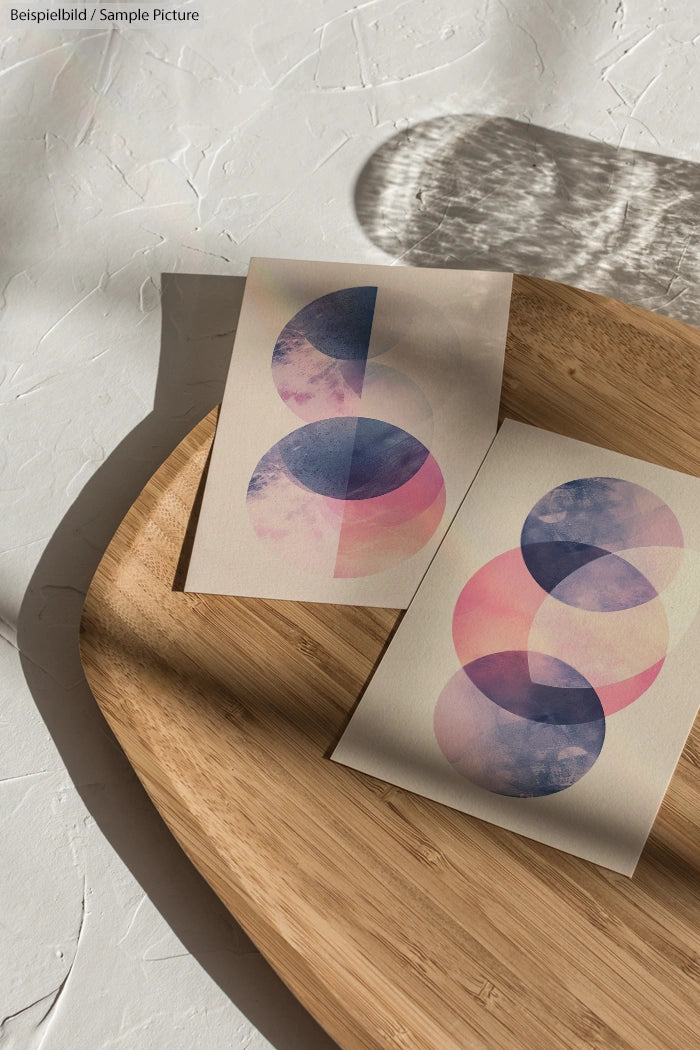 Two art prints with overlapping circular patterns in blue and pink hues on a wooden table with shadows cast on a textured surface.