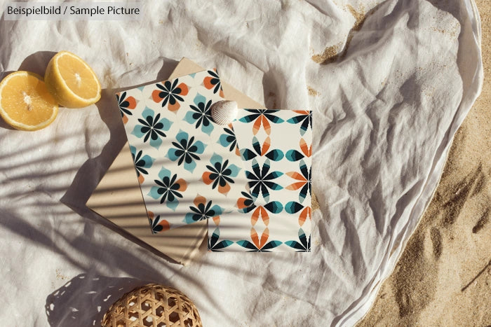 Beach scene with patterned notebooks, sliced lemon, and woven accessory on sand and white fabric.