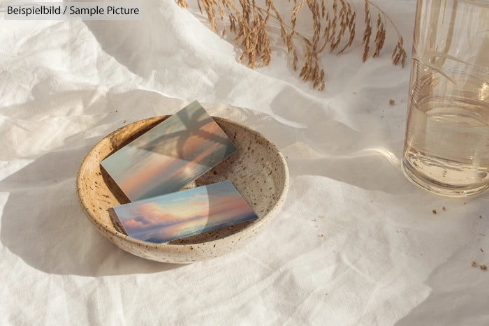 Ceramic bowl with artistic cards on white cloth next to a glass of water and dry grass.