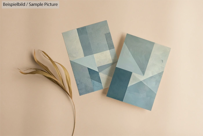 Two abstract geometric art pieces with blue tones on a beige background, next to a dried plant stem.