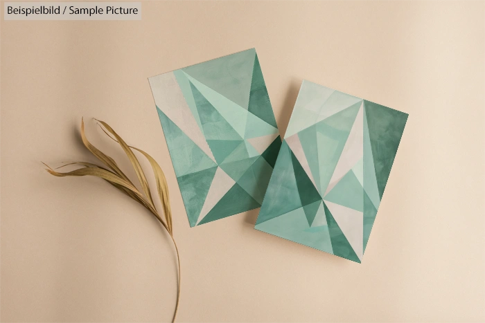 Two abstract geometric paintings in shades of teal on a beige surface with dried grass beside them.