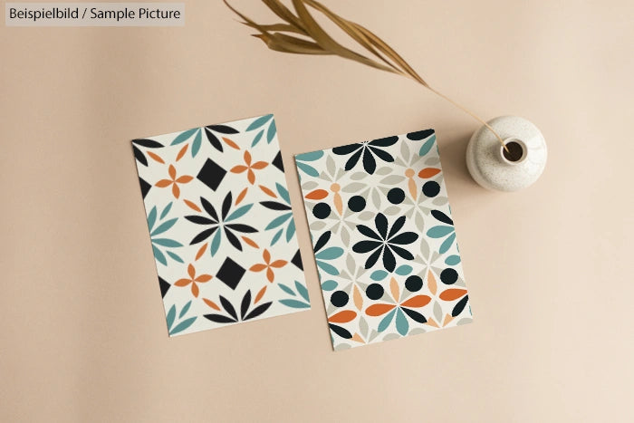 Two geometric pattern postcards with floral designs on a beige surface beside a ceramic vase with dried flowers.