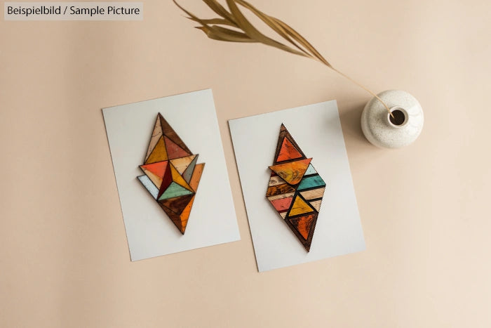 Two geometric mosaic artworks on paper next to a vase with dried grass on a neutral background.