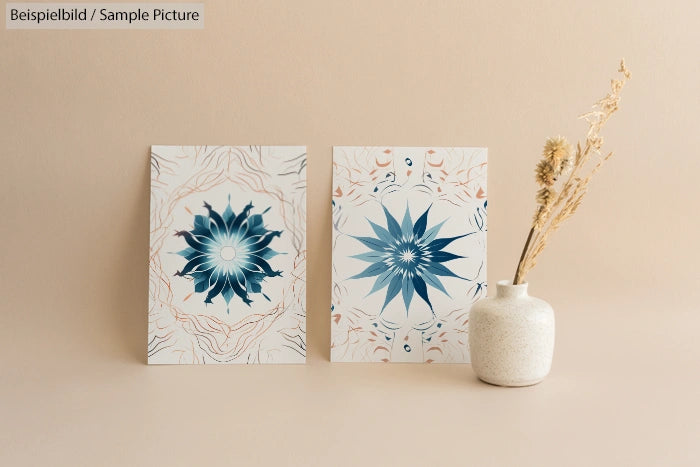 Two decorative prints with blue floral designs beside a white ceramic vase with dried flowers on a beige background.