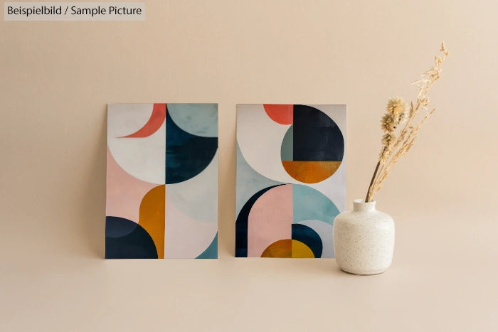 Two abstract geometric paintings with earthy tones next to a white vase on a beige background.