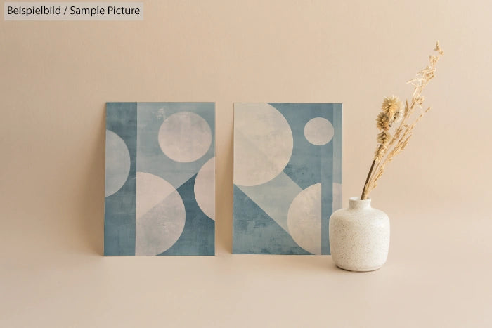 Two abstract blue and white geometric prints next to a white vase with dried flowers on a beige background.