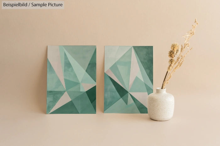 Two geometric abstract paintings in teal tones beside a white vase with dried plants on a beige background.