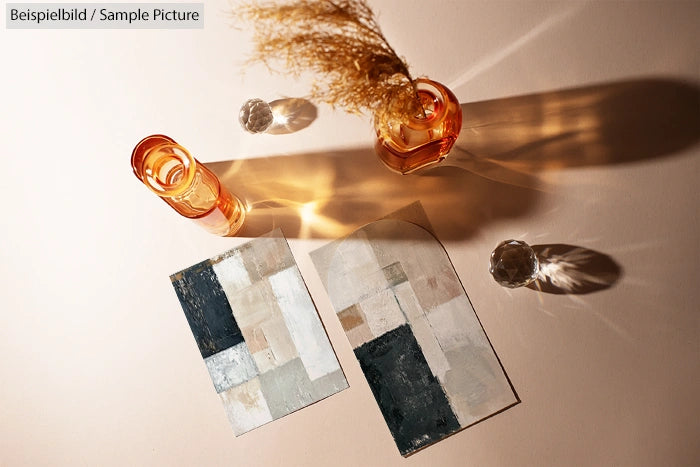 Abstract geometric paintings on a table with amber glassware casting shadows. Dried grass in a vase.